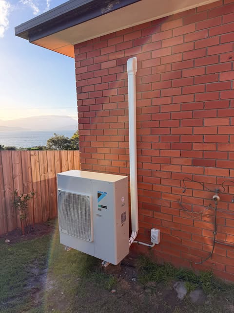 Split system heat pump outdoor unit on a brick wall with its isolator switch and conduit